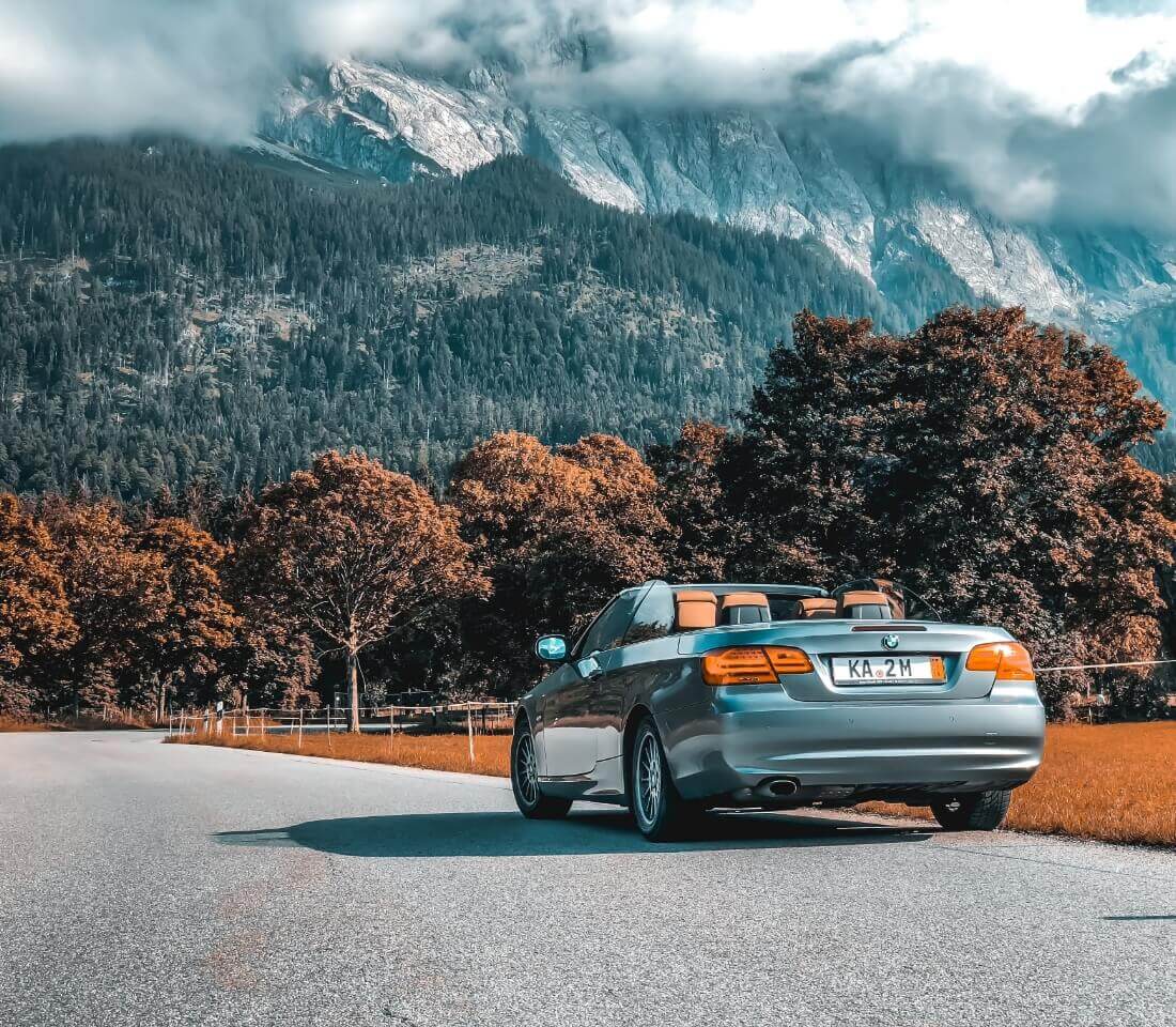 山々と青空を背景に、木々に囲まれた道を走る銀色のオープンカーの後方の風景