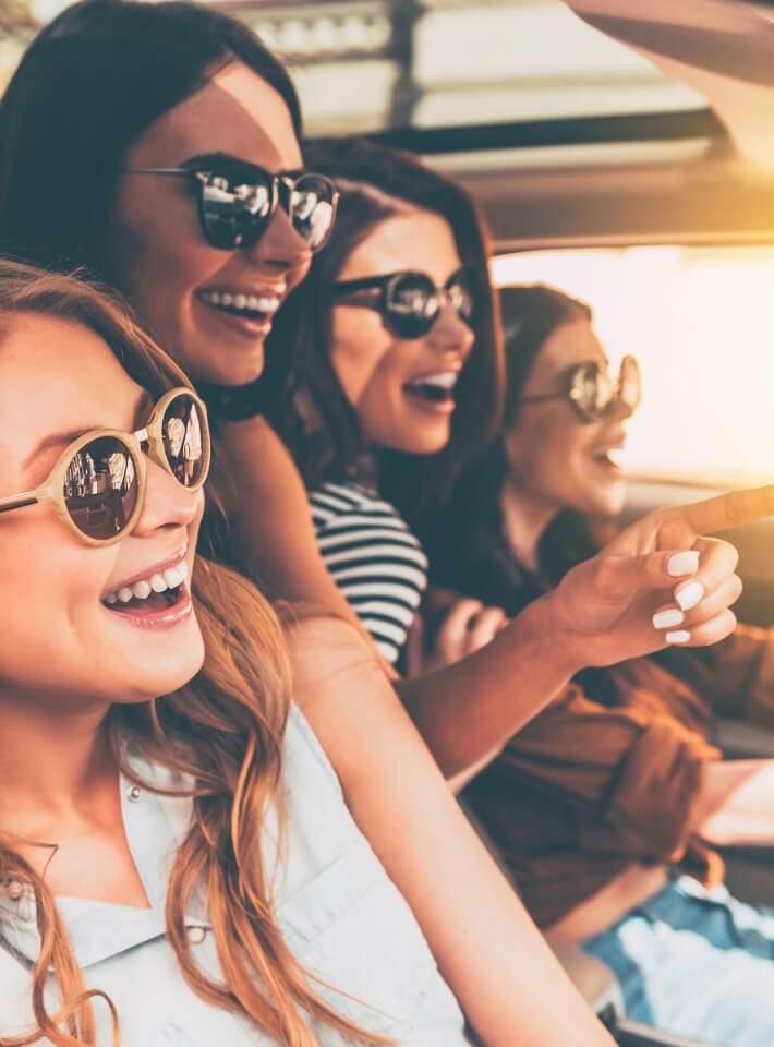 若い女性たちが車内でサングラスをかけて笑い合い、楽しそうにドライブしている様子。