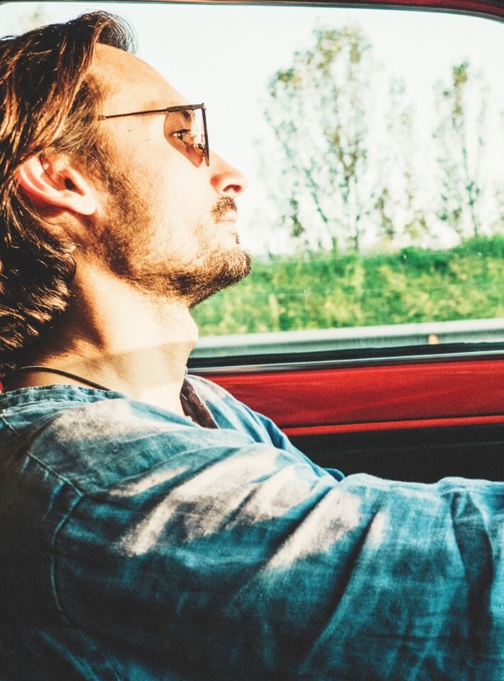 サングラスをかけた男性が赤い車を運転し、リラックスしながら風景を眺めているシーン。