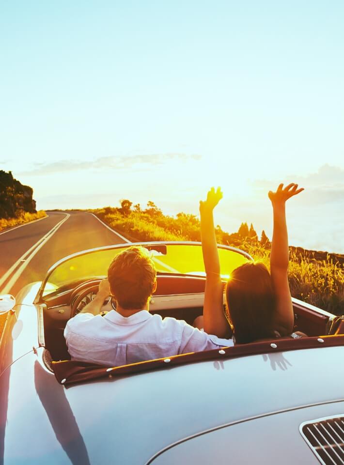 オープンカーに乗った二人が、夕日に向かって手を挙げながら爽やかな景色を満喫している場面。