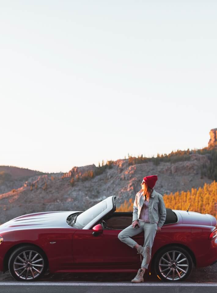 赤いオープンカーの横に立つ女性が、帽子とカジュアルな服装で夕焼けの景色を背景にポーズをとっている姿。